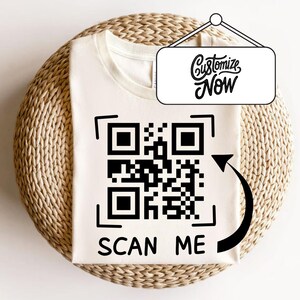 May include: Cream-coloured t-shirt featuring a large black QR code and the words "SCAN ME" printed below. A black arrow curves around the QR code. A sign above the shirt reads "Customize Now" in a script font.