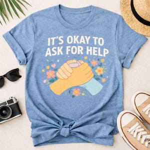 May include: Pale blue t-shirt with the white text "IT'S OKAY TO ASK FOR HELP". The graphic shows two clasped hands, with floral accents. The t-shirt is knotted at the waist.