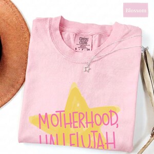 K&ouml;nnte beinhalten: Ein hellrosa T-Shirt mit einer gro&szlig;en gelben Sterngrafik und dem Text "MOTHERHOOD, HALLELUJAH" in rosa Schrift. Das T-Shirt hat ein Comfort Colors Markenetikett am inneren Halsausschnitt und eine zarte silberfarbene Halskette mit Sternanh&auml;nger.