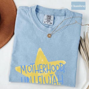 K&ouml;nnte beinhalten: Ein hellblaues T-Shirt mit Rundhalsausschnitt und einer gro&szlig;en gelben Stern-Grafik auf der Vorderseite. Der Text "MOTHERHOOD, HALLELUJAH" ist in Blau &uuml;ber den Stern gedruckt. Das Shirt hat ein Comfort Colors Etikett am Hals und eine goldfarbene Kette.