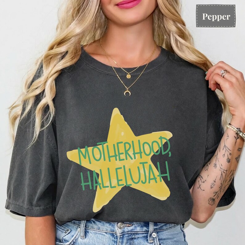 K&ouml;nnte beinhalten: Ein anthrazitfarbenes Kurzarm-T-Shirt mit einem gro&szlig;en gelben Stern-Grafikmotiv in der Mitte der Brust. Der Text "MOTHERHOOD, HALLELUJAH" ist in gr&uuml;nen Gro&szlig;buchstaben &uuml;ber den Stern gedruckt. Das T-Shirt hat einen Rundhalsausschnitt und eine lockere Passform.