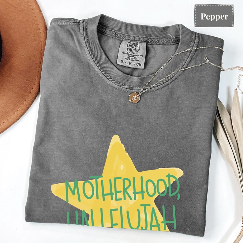K&ouml;nnte beinhalten: Ein pfefferfarbenes T-Shirt mit einer gro&szlig;en gelben Stern-Grafik und dem gr&uuml;nen Schriftzug MOTHERHOOD, HALLELUJAH. Das T-Shirt hat einen Rundhalsausschnitt und ein Comfort Colors Markenetikett am Innenkragen. Eine d&uuml;nne silberne Halskette liegt auf dem Stoff.