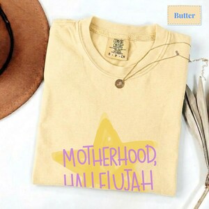 K&ouml;nnte beinhalten: Ein buttergelbes Kurzarm-T-Shirt mit einer gro&szlig;en, hellgelben Sternengrafik auf der Vorderseite. Der Text "MOTHERHOOD, HALLELUJAH" ist in Lila &uuml;ber den Stern gedruckt. Das T-Shirt hat einen Rundhalsausschnitt und ein Comfort Colors Markenetikett.