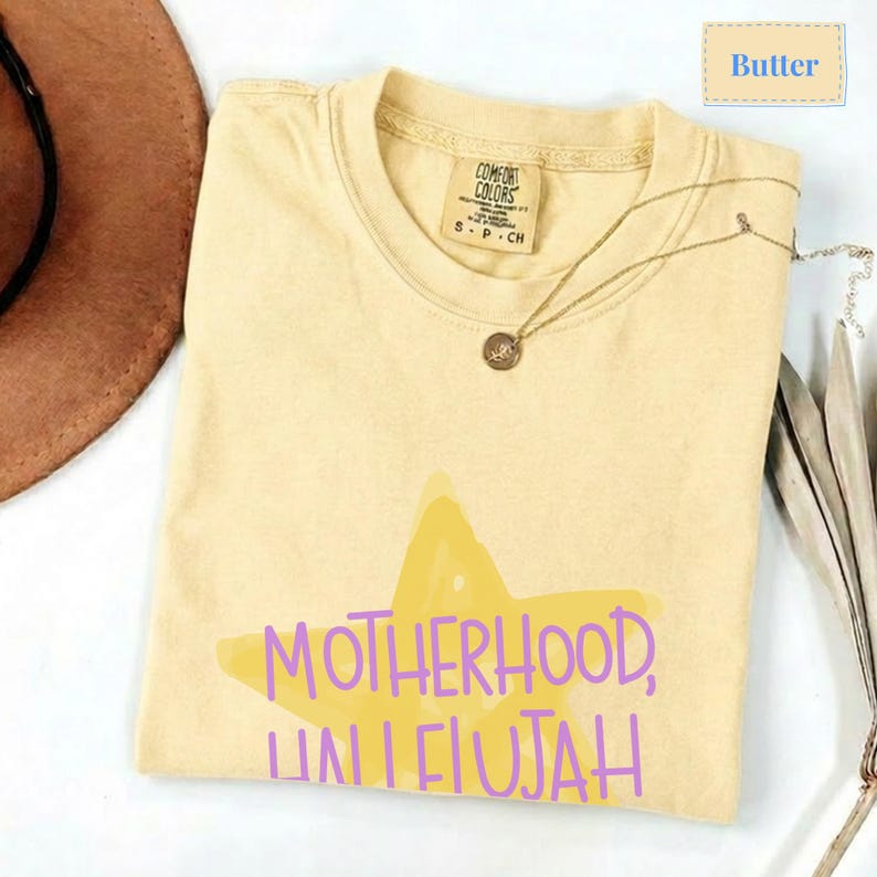 K&ouml;nnte beinhalten: Ein buttergelbes Kurzarm-T-Shirt mit einer gro&szlig;en, hellgelben Sternengrafik auf der Vorderseite. Der Text "MOTHERHOOD, HALLELUJAH" ist in Lila &uuml;ber den Stern gedruckt. Das T-Shirt hat einen Rundhalsausschnitt und ein Comfort Colors Markenetikett.