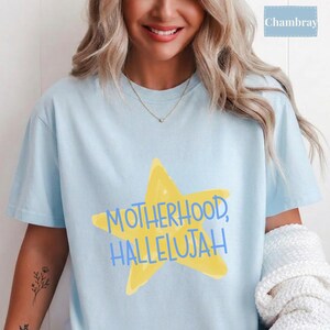 K&ouml;nnte beinhalten: Ein hellblaues Kurzarm-T-Shirt mit einer gro&szlig;en gelben Stern-Grafik in der Mitte der Brust. Der Text "MOTHERHOOD, HALLELUJAH" ist in blauen Gro&szlig;buchstaben &uuml;ber den Stern gedruckt. Das T-Shirt hat einen Rundhalsausschnitt und eine lockere Passform.