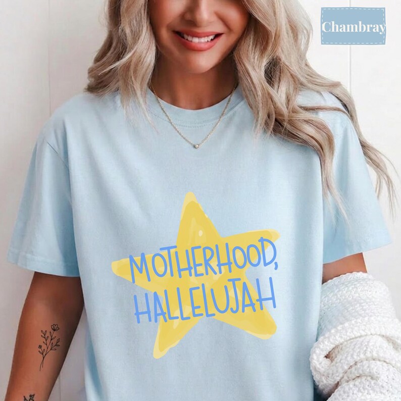 K&ouml;nnte beinhalten: Ein hellblaues Kurzarm-T-Shirt mit einer gro&szlig;en gelben Stern-Grafik in der Mitte der Brust. Der Text "MOTHERHOOD, HALLELUJAH" ist in blauen Gro&szlig;buchstaben &uuml;ber den Stern gedruckt. Das T-Shirt hat einen Rundhalsausschnitt und eine lockere Passform.
