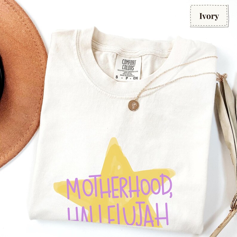 K&ouml;nnte beinhalten: Ein elfenbeinfarbenes T-Shirt mit kurzen &Auml;rmeln, einem gelben Stern-Motiv und dem violetten Schriftzug "MOTHERHOOD, HALLELUJAH". Das Shirt hat einen Rundhalsausschnitt, ein Comfort Colors Markenetikett am Innenkragen und eine goldene Halskette.