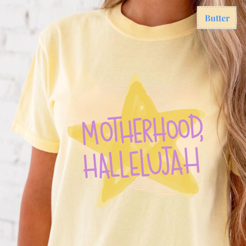 K&ouml;nnte beinhalten: Ein buttergelbes T-Shirt mit kurzen &Auml;rmeln und einer gro&szlig;en, hellgelben Sternengrafik in der Mitte der Brust. &Uuml;ber dem Stern befindet sich der violette Schriftzug "MOTHERHOOD, HALLELUJAH" in einer lockeren, handschriftlichen Schriftart.