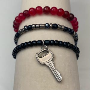 May include: Three beaded bracelets. The top bracelet has red beads. The middle bracelet has dark blue and silver beads. The bottom bracelet has black beads and a silver key charm.