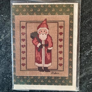 Pode incluir: Um cartão de Natal com um desenho de ponto cruz do Pai Natal com um saco. O cartão tem um Pai Natal vermelho e branco, um fundo castanho e uma borda verde com um padrão repetido. O nome "Robin" está impresso no cartão.