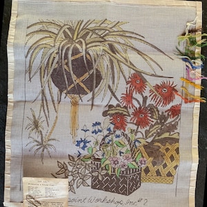 May include: A needlepoint canvas featuring a hanging plant in a dark brown pot, a basket of red flowers, and a smaller basket of blue and pink flowers. The canvas includes a color key label and loose yarn strands attached to the right side edge.
