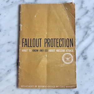 Puede incluir: Un folleto vintage de color beige con el título "FALLOUT PROTECTION" y el subtítulo "WHAT TO KNOW AND DO ABOUT NUCLEAR ATTACK". La portada presenta texto negro y un sello del Departamento de Defensa. El folleto está desgastado.