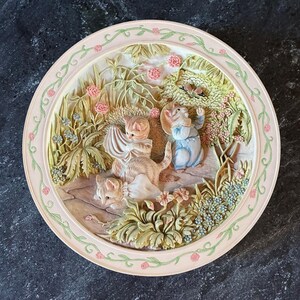 May include: A round decorative wall plate featuring a three-dimensional relief scene of two orange tabby kittens and a rabbit wearing blue overalls in a garden setting with pink flowers, green foliage, and a butterfly, framed by a floral border.