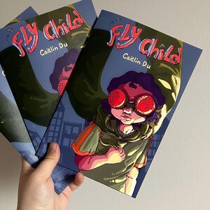 May include: A stack of comic books titled "Fly Child" by Caitlin Du. The cover art features a child with red-tinted goggles and a green cape against a blue cityscape background. The title is in pink and red.