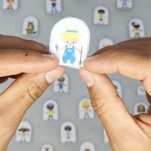 Op de afbeelding: Een sticker met een cartoonillustratie van een persoon in een blauwe overall met een schop en een troffel. De sticker wordt tussen twee handen gehouden, met andere soortgelijke stickers op de achtergrond.