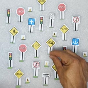 Op de afbeelding: Een verzameling miniatuur verkeersborden, waaronder "STOP", "SLOW" en "SPEED LIMIT 50", samen met verkeerslichten en andere symbolen. De borden zijn wit met verschillende gekleurde randen en iconen, gerangschikt op een lichtgrijze ondergrond.