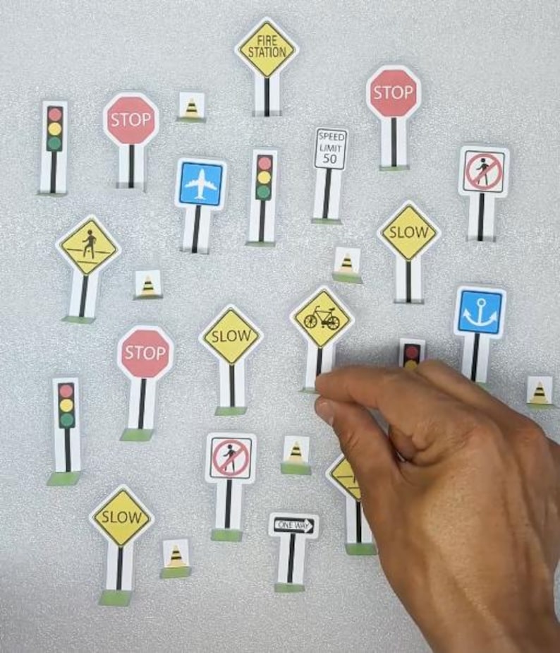 Op de afbeelding: Een verzameling miniatuur verkeersborden, waaronder "STOP", "SLOW" en "SPEED LIMIT 50", samen met verkeerslichten en andere symbolen. De borden zijn wit met verschillende gekleurde randen en iconen, gerangschikt op een lichtgrijze ondergrond.