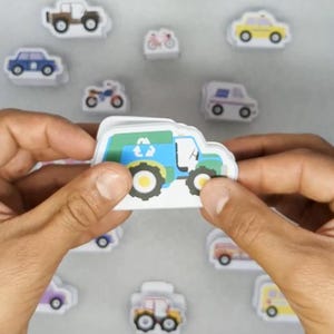 Op de afbeelding: Een persoon houdt een recyclingvrachtwagensticker vast met een blauw, groen en wit ontwerp. Rondom de vrachtwagen bevinden zich diverse voertuigstickers, waaronder auto's, ambulances en motorfietsen. De stickers zijn wit met kleurrijke illustraties.