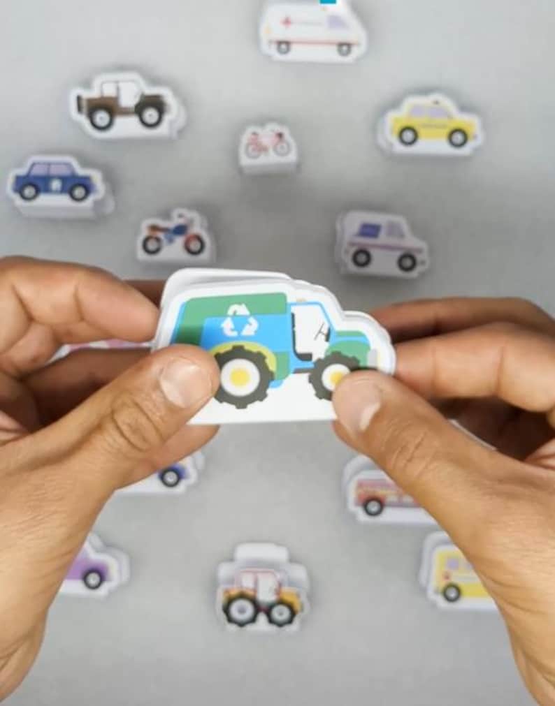 Op de afbeelding: Een persoon houdt een recyclingvrachtwagensticker vast met een blauw, groen en wit ontwerp. Rondom de vrachtwagen bevinden zich diverse voertuigstickers, waaronder auto's, ambulances en motorfietsen. De stickers zijn wit met kleurrijke illustraties.