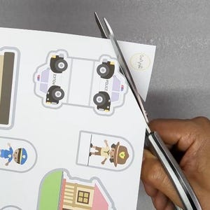 Op de afbeelding: Schaar die een vel papier knipt met kleurrijke illustraties van politieauto's, gebouwen en mensen. De illustraties zijn in cartoonstijl, met heldere kleuren en eenvoudige ontwerpen. Het papier is wit en de schaar is zilverkleurig.