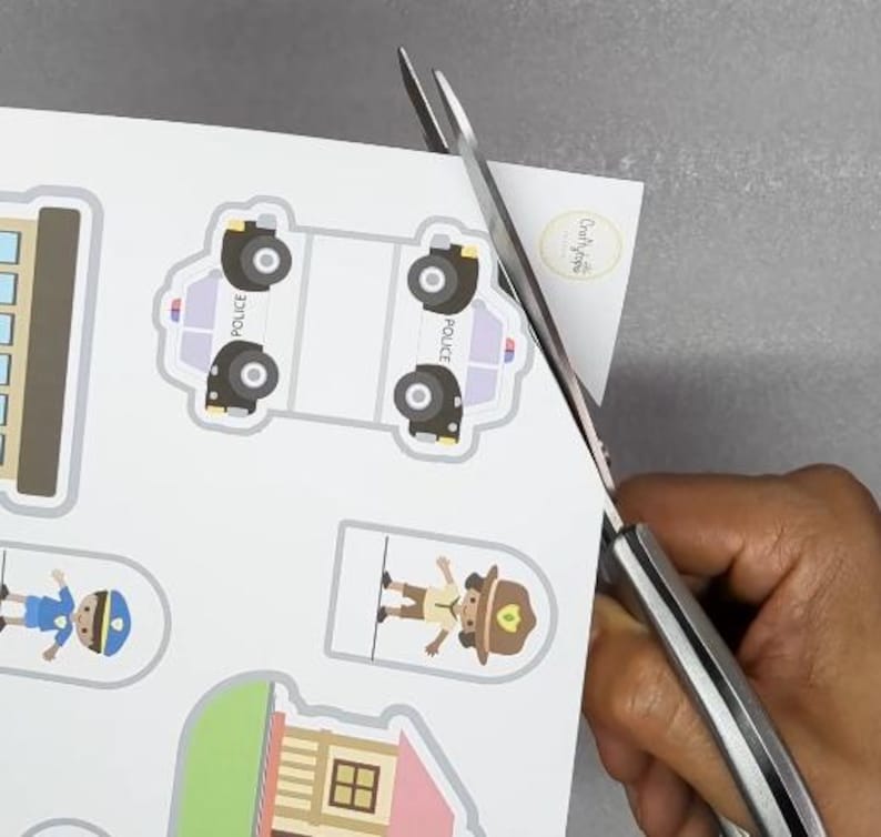 Op de afbeelding: Schaar die een vel papier knipt met kleurrijke illustraties van politieauto's, gebouwen en mensen. De illustraties zijn in cartoonstijl, met heldere kleuren en eenvoudige ontwerpen. Het papier is wit en de schaar is zilverkleurig.