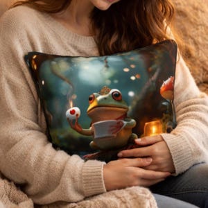 May include: Square cushion with a fantasy frog character holding a teacup and a heart-shaped treat. The frog is set against a forest backdrop with soft lighting. The cushion is held by a person wearing a beige jumper.
