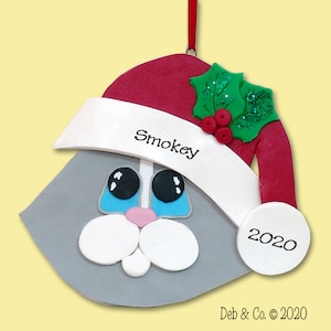 May include: A gray cat-shaped Christmas ornament wearing a red Santa hat with a white brim. The ornament has black eyes, a pink nose, and white whiskers. The name "Smokey" is written on the hat, and the year "2020" is on a white circle.