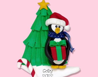 May include: A penguin wearing a red Santa hat and blue scarf holds a green wrapped gift in front of a green Christmas tree. The ornament is personalized with the name "Colby" and the year "2022".