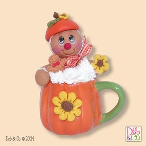 May include: A whimsical ceramic ornament featuring a gingerbread person in a pumpkin-shaped mug. The gingerbread person wears an orange hat and holds a small gingerbread cookie. The mug is orange with a green handle and yellow flower accents.