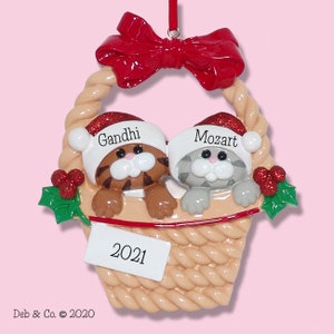 May include: A Christmas ornament featuring two cat figurines in a woven basket. The cats wear Santa hats with the names "Gandhi" and "Mozart." A red bow tops the basket, with "2021" on a tag. The ornament includes holly accents.