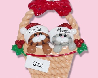 May include: A Christmas ornament featuring two cat figurines in a woven basket. The cats wear Santa hats with the names "Gandhi" and "Mozart." A red bow tops the basket, with "2021" on a tag. The ornament includes holly accents.
