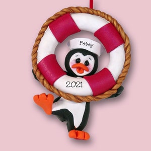 May include: A handmade penguin Christmas ornament wearing a sailor hat and holding a red and white life preserver. The ornament is decorated with the name "Petey" and the year "2021".