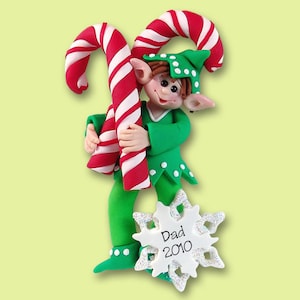 May include: A green elf ornament holding two candy canes. The elf is wearing a green hat with white polka dots and a green and white outfit. The ornament has a white snowflake with the text "Dad 2010" on it.