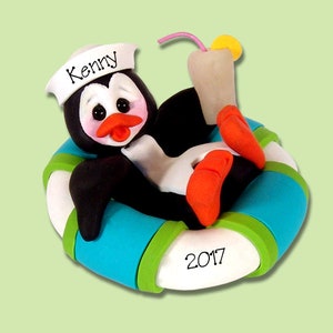 May include: A penguin wearing a sailor hat and holding a drink floats in a blue and green inner tube. The inner tube has the name "Kenny" and the year "2017" written on it.