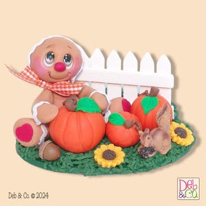 May include: A whimsical autumn-themed figurine featuring a gingerbread person with a scarf, sitting next to orange pumpkins, sunflowers, and a squirrel. A white picket fence is in the background, all on a green base. The figurine has a heart on its knee.