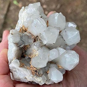 May include: A cluster of clear quartz crystals with multiple facets. The crystals are a translucent white color, with some areas showing a slight gray hue. The quartz is set against a backdrop of a light brown matrix.