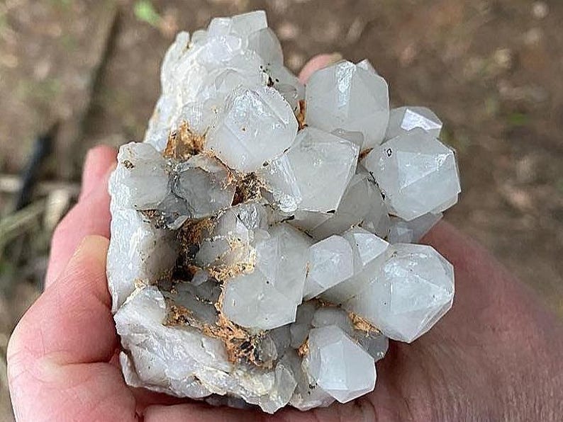 May include: A cluster of clear quartz crystals with multiple facets. The crystals are a translucent white color, with some areas showing a slight gray hue. The quartz is set against a backdrop of a light brown matrix.