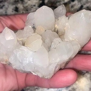 May include: A cluster of clear quartz crystals with a variety of shapes and sizes. The crystals are held in a hand, showcasing their natural, translucent appearance. The quartz has a slightly milky hue.