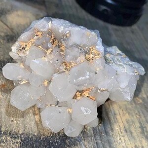 May include: A cluster of clear quartz crystals with a crystalline structure. The quartz has a translucent appearance with hints of gold and is set against a wooden surface. The crystals vary in size and shape, creating a natural, organic aesthetic.
