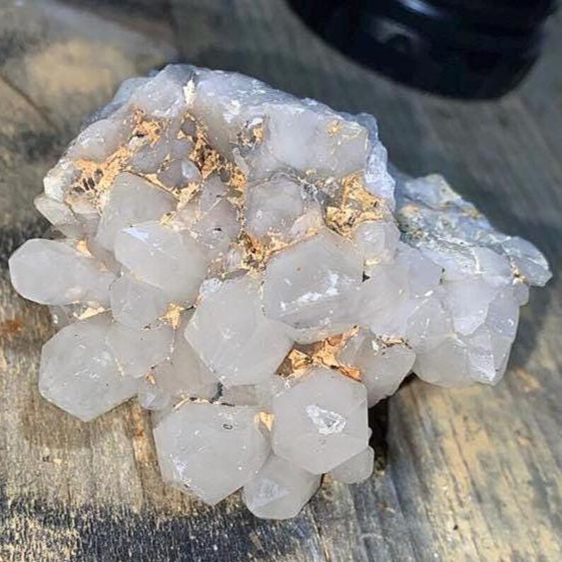May include: A cluster of clear quartz crystals with a crystalline structure. The quartz has a translucent appearance with hints of gold and is set against a wooden surface. The crystals vary in size and shape, creating a natural, organic aesthetic.