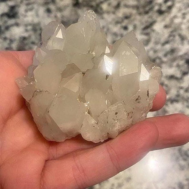 May include: A cluster of clear quartz crystals with multiple points. The crystals are held in a hand, showcasing their natural, irregular shape and translucent quality. The quartz has a slightly milky appearance.