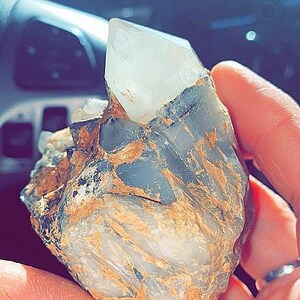 May include: A clear quartz crystal point emerges from a rough, multi-colored rock formation. The rock displays shades of gray, brown, and tan, with a textured surface. The crystal is translucent and sharply pointed.