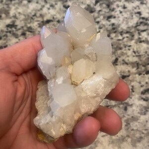 May include: A cluster of clear quartz crystals with multiple points. The crystals are held in a hand, showcasing their natural, irregular shape and translucent quality. The quartz has a pale, off-white color.