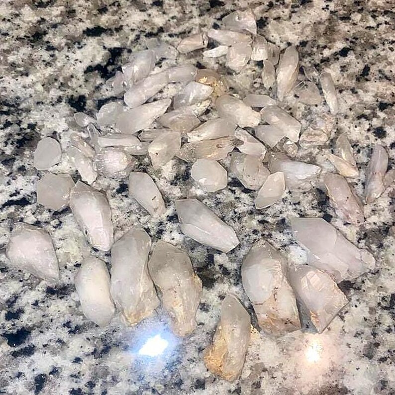 May include: A collection of clear quartz crystal points, varying in size and shape. The crystals are translucent with some having a slightly milky appearance. They are scattered on a speckled, gray and white surface.