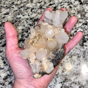 May include: A cluster of clear quartz crystals with a variety of shapes and sizes, held in a hand. The crystals have a translucent appearance with some areas of light brown and are attached to a dark, rocky base. The background is a speckled gray surface.