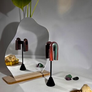 May include: A pair of abstract earrings with a red, green, and brown color scheme. The earrings are displayed on a black stand in front of a wavy mirror. Several small, colorful stones are scattered around the base of the stand.