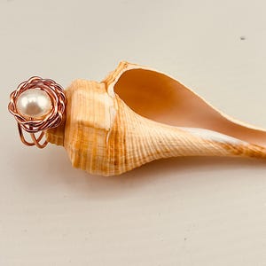 May include: A copper-colored wire ring with a large, round, white pearl center. The ring is next to a large, tan seashell with a smooth, pearlescent interior. The seashell is resting on a light-colored surface.