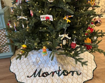 May include: A round, quilted Christmas tree skirt with a scalloped edge, featuring a white fabric base with a gold-toned lattice pattern. The name Morton is appliqued in the center in dark green fabric with a red and white floral print.
