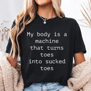 May include: Black t-shirt with the text "My body is a machine that turns toes into sucked toes" in white. The shirt is a crew neck style.