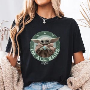 May include: Black t-shirt featuring a graphic of Grogu, also known as Baby Yoda, from The Mandalorian. The design includes the text "I'M ALL EARS" in green around Grogu. The shirt is a crew neck style.